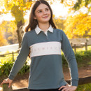LeMieux long-sleeve equestrian polo top in muted green with white collar and branded white chest panel, worn outdoors beside a wooden fence.