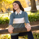 Lifestyle view of a young rider wearing a green LeMieux long-sleeve polo top with white branded chest panel beside a wooden fence in an autumn setting.