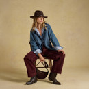 Model sitting on a chair, wearing a denim jacket, striped shirt, burgundy pants, and brown cowboy boots, holding a bag.