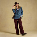 A woman in a denim shirt and maroon pants poses against a neutral background, wearing a brown hat and carrying a bag.