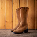 Brown suede Western-style boots with a stacked heel on a textured floor against a wooden background.