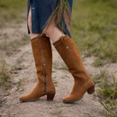 A pair of tall chestnut brown suede boots with a block heel, worn by a person standing on a dirt path.
