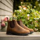 A pair of brown Chelsea boots with elastic side panels, placed on a wooden surface with a blurred garden background.