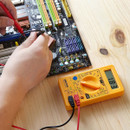 A DEKTON digital multimeter with measurement leads rests on a wooden surface next to a computer motherboard.
