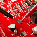 Close-up of a red circuit board with a microchip, multimeter probes measuring voltage on the chip.