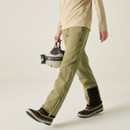 A person is walking while wearing martolive green trousers and holding a grey and black helmet.