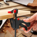 A hand is using a black and red clamp on a wooden board, suitable for holding materials together.