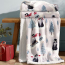 White sherpa fleece Christmas throw featuring playful penguins, winter trees and festive motifs, draped over a wooden bench.