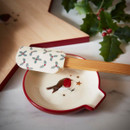A round cream ceramic spoon rest with a red rim, featuring a robin design, alongside a patterned silicone spatula with a bamboo handle.