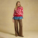 A woman stands wearing a red knitted jumper with white sheep, layered over a denim shirt, paired with brown checked wide-leg trousers.