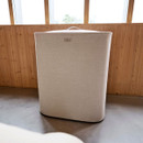 A soft-edged, beige laundry basket with a textured fabric exterior, positioned against a wooden wall with large windows.
