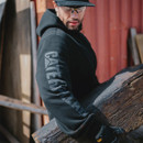 A man wearing a black hooded sweatshirt with a large logo on the sleeve, holding a wooden beam outdoors.