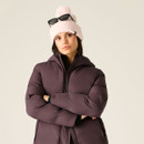 A woman poses in a puffy, dark purple jacket with a high collar, wearing a light pink beanie and black sunglasses.