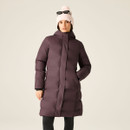 A woman is wearing a long, puffy, burgundy jacket with a hood, paired with a pink beanie and sunglasses, against a neutral background.
