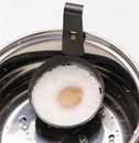 Black non-stick egg poacher cup with a metal hanging hook, holding a cooked poached egg with a pale yellow yolk, positioned inside a stainless steel saucepan with water droplets visible.