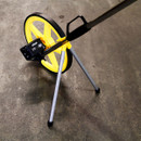 Yellow and black measuring wheel with a stand, resting on a concrete surface. The wheel is partially folded, conveying utility and functionality.