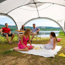 A family enjoying a picnic under a large canopy by a lake, with adults seated at a table and two children playing on a blanket.
