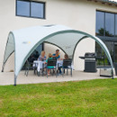 A family gathers under a large white canopy, enjoying a meal at a table with a barbecue grill nearby, set against a house backdrop.