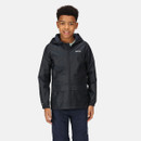 A young boy smiles while wearing a black hooded rain jacket and dark pants against a plain light gray background, conveying a cheerful mood.