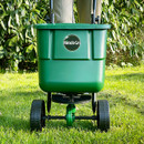 Green fertilizer spreader labeled "Miracle-Gro" on a lawn. Two wheels, standing upright, with blurred greenery in the background, conveying outdoor use.