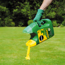 A person wearing green gloves holds a green and yellow garden sprayer over a lush lawn. The background features blurred greenery and trees.
