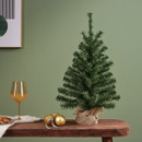 Mini artificial Christmas tree on a wooden table with festive decorations, showcasing dense evergreen foliage and a natural-look burlap pot cover.