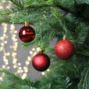 Red Christmas baubles hanging on a fir tree, featuring glossy, matte and glitter finishes with warm white fairy lights in the background.