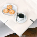White tablecloth secured to a dining table with silver metal clips, shown alongside a white plate holding three bread rolls and a cup of black coffee on a saucer.