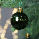 Glossy dark green Christmas bauble hanging from an evergreen branch, reflecting festive lights and seasonal surroundings.