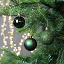 Dark green Christmas baubles hanging on a fir tree, featuring glossy, matte and glitter finishes with warm white fairy lights in the background.