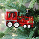 Decorative red locomotive Christmas tree ornament with glitter finish, black chimney and festive wreath accents, displayed on evergreen branches.