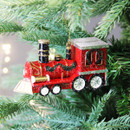 Red steam train Christmas ornament with sparkling finish, festive greenery trim and silver glitter roof, suspended on a Christmas tree branch.