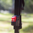 Close-up of a red button on a black device mounted on a pole, set against a blurred green outdoor background.