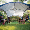 A group of people relaxes under a large canopy in a grassy area surrounded by trees, enjoying a sunny outdoor gathering.