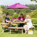 Three people sitting at a round wooden picnic table with built-in benches under a pink parasol in a garden.