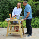 Two people standing beside a wooden BBQ side table on a patio, preparing food outdoors.