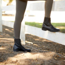 Close-up of a person wearing black riding boots and beige leggings, standing on a dusty ground beside a white wooden fence, suggesting an equestrian setting.
