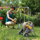 Hozelock wheeled hose reel cart in use in a garden, with a woman unwinding the hose to water plants.