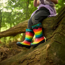 Child sitting on a tree branch, wearing colourful rainbow-striped rain boots and dark jeans, surrounded by lush green forest foliage. Playful and adventurous mood.