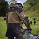 A person in a muddy, brown and black jacket stands beside an ATV in a grassy field with grazing cattle, wearing a white helmet, evoking a rural, adventurous feel.