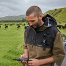 A man in a brown jacket uses a handheld device in a lush green pasture with grazing cows. The scenery includes rolling hills under a cloudy sky, conveying a serene rural setting.