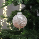 A blush pink ombre, sequinned bauble hanging from a Christmas tree branch, with gold string lights in the background.