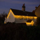 Traditional cottage decorated with warm white LED snowing icicle lights along the roofline and porch, creating a festive outdoor Christmas display at dusk.