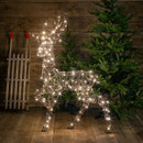 Illuminated wireframe stag Christmas decoration wrapped in warm white LED lights, displayed indoors beside fir trees for a festive seasonal display.