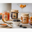 Open jars of Edinburgh Preserves Gin Preserves on a table with fresh fruit, gold spoon, and a cocktail glass. The jams include Apricot and Passionfruit, Blackcurrant, and Pink Grapefruit and Mandarin.