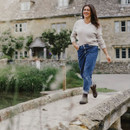 Woman in a beige jumper and jeans walks joyfully on a stone bridge. Background shows an ivy-covered stone cottage and lush greenery, conveying a serene mood.
