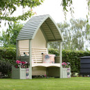 Wide garden view of sage green painted arbour bench with slatted back and curved roof, flanked by sage planters on a lawn.