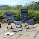 Two navy blue reclining chairs sit on a stone patio, surrounded by greenery and hills, with a magazine and wine glass nearby.