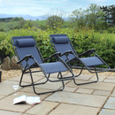 Two navy blue reclining chairs positioned on a patio, surrounded by lush greenery and distant hills, with a magazine and glass nearby.