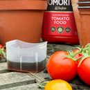 Small plastic measuring cup with dark liquid, surrounded by ripe red and yellow tomatoes, terracotta pots, and a bottle of concentrated tomato food. Gardening scene.